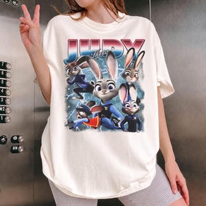 Op de afbeelding: Cr&egrave;mekleurig T-shirt met een afbeelding van Judy Hopps en andere personages uit de animatiefilm Zootopia. De afbeelding bevat het woord "JUDY" in een retro lettertype met een blauwe en rode gradi&euml;nt. Het shirt heeft een losse pasvorm.