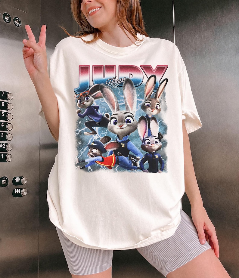 Op de afbeelding: Cr&egrave;mekleurig T-shirt met een afbeelding van Judy Hopps en andere personages uit de animatiefilm Zootopia. De afbeelding bevat het woord "JUDY" in een retro lettertype met een blauwe en rode gradi&euml;nt. Het shirt heeft een losse pasvorm.