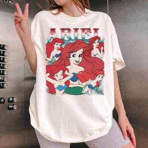 Pu&ograve; includere: T-shirt color crema con grafica di Ariel de La Sirenetta. Il design include la scritta "ARIEL" in rosso e diverse illustrazioni di Ariel con capelli rossi e reggiseno a conchiglia blu. La maglietta &egrave; di vestibilit&agrave; ampia.