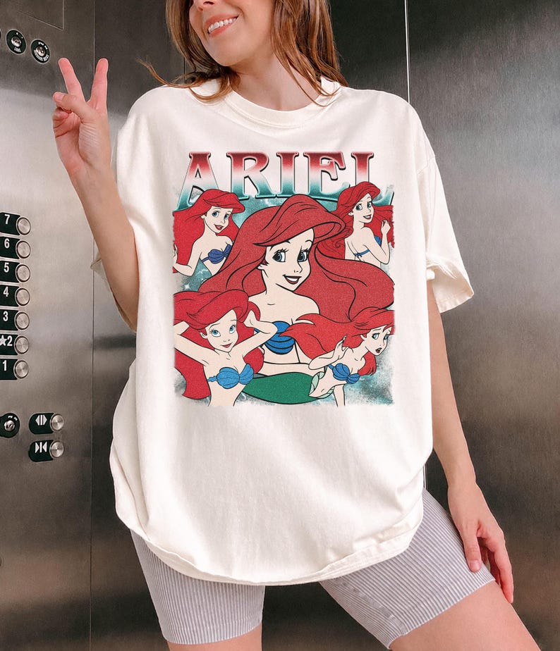 Pu&ograve; includere: T-shirt color crema con grafica di Ariel de La Sirenetta. Il design include la scritta "ARIEL" in rosso e diverse illustrazioni di Ariel con capelli rossi e reggiseno a conchiglia blu. La maglietta &egrave; di vestibilit&agrave; ampia.