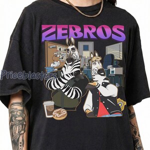 Pu&ograve; includere: T-shirt grigio scuro con un'illustrazione a cartoni animati di due zebre in un ufficio. Il testo "ZEBROS" &egrave; visualizzato sopra le zebre in una sfumatura di colore viola e rosa. Una zebra indossa una giacca e l'altra un gilet.