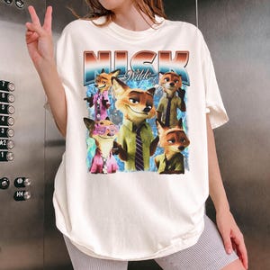 Pode incluir: Camiseta creme com um gr&aacute;fico de Nick Wilde de Zootopia. O gr&aacute;fico inclui o nome "Nick Wilde" em uma fonte retr&ocirc;, junto com v&aacute;rias imagens do personagem. A camiseta tem um caimento folgado.