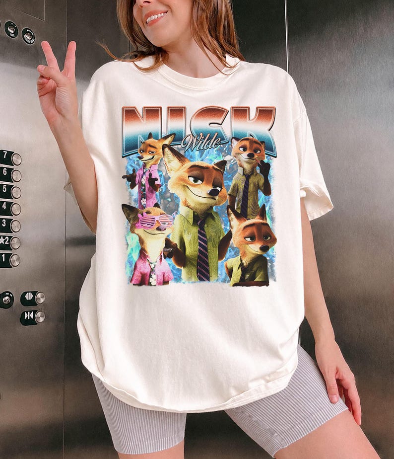 Pode incluir: Camiseta creme com um gr&aacute;fico de Nick Wilde de Zootopia. O gr&aacute;fico inclui o nome "Nick Wilde" em uma fonte retr&ocirc;, junto com v&aacute;rias imagens do personagem. A camiseta tem um caimento folgado.