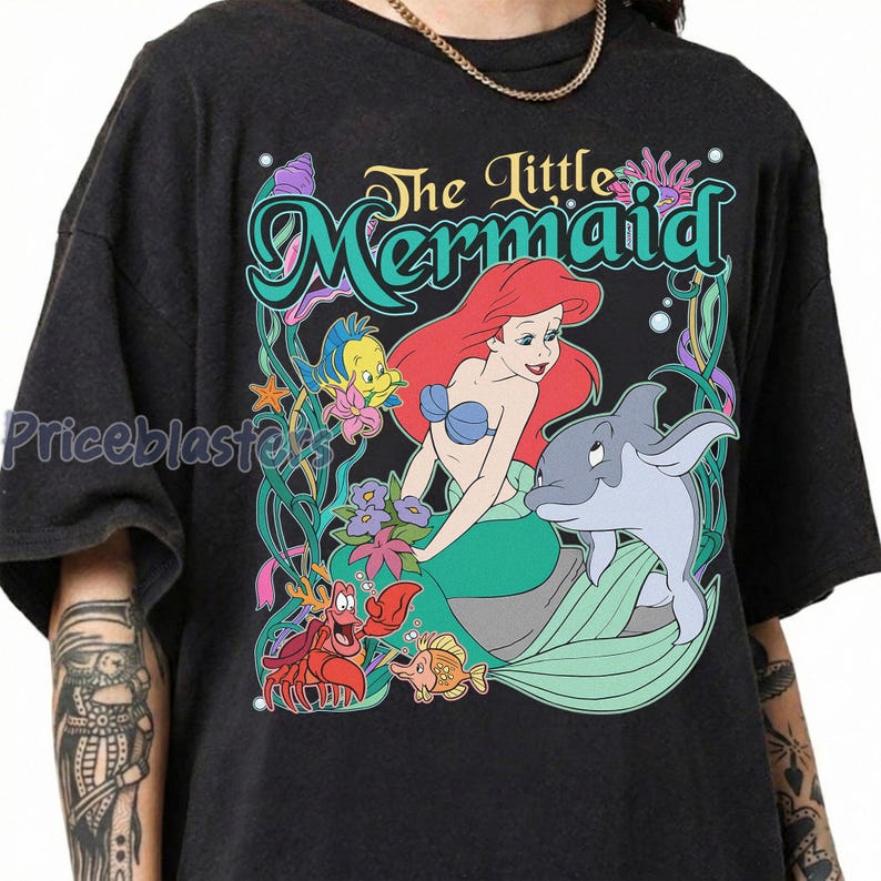 May include: Dark gray t-shirt featuring a colorful graphic of Ariel from The Little Mermaid. The design includes Ariel, a dolphin, and other sea creatures, with the words "The Little Mermaid" in a decorative font.