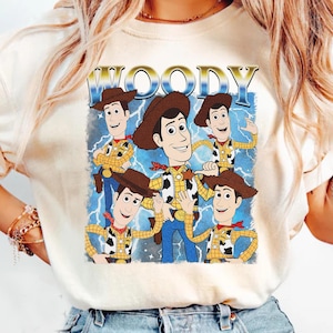 May include: Cream-colored t-shirt featuring a graphic of Woody from Toy Story. The design includes multiple images of Woody in his cowboy attire, with the word "WOODY" in a gold, retro font. Blue lightning bolts are in the background.