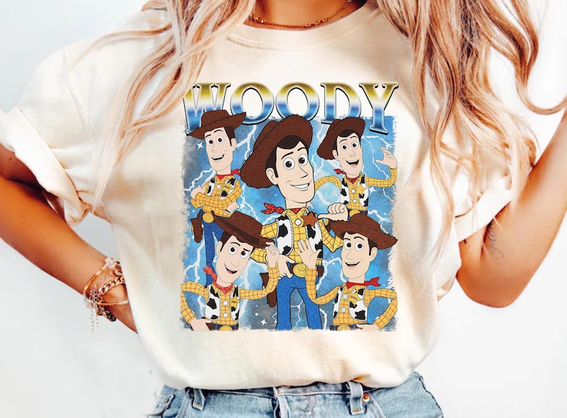 May include: Cream-colored t-shirt featuring a graphic of Woody from Toy Story. The design includes multiple images of Woody in his cowboy attire, with the word "WOODY" in a gold, retro font. Blue lightning bolts are in the background.