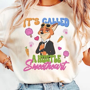 May include: Cream-colored t-shirt featuring a cartoon fox in a suit and sunglasses. The shirt has the text "IT'S CALLED A HUSTLE Sweetheart" in colorful lettering, along with paw prints, carrots, and hearts.