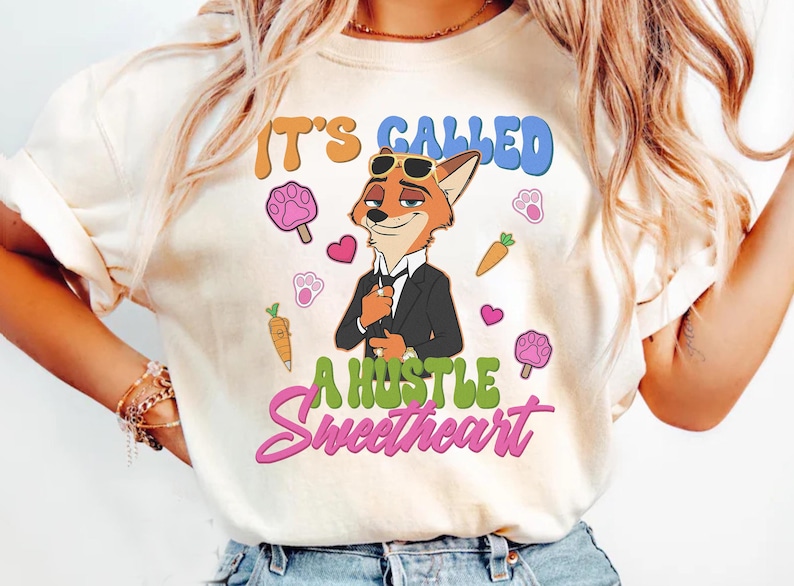 May include: Cream-colored t-shirt featuring a cartoon fox in a suit and sunglasses. The shirt has the text "IT'S CALLED A HUSTLE Sweetheart" in colorful lettering, along with paw prints, carrots, and hearts.