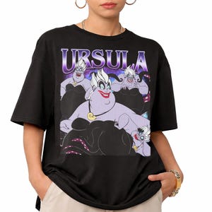 May include: Black t-shirt featuring a graphic of Ursula from The Little Mermaid. The design includes the word "URSULA" in a purple font above an illustration of Ursula in various poses. The shirt is a short-sleeved crew neck.