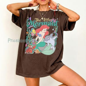 May include: Brown t-shirt featuring a graphic of Ariel from The Little Mermaid. The design includes Ariel with red hair, a blue top, and a green tail, along with a dolphin and other sea creatures. The text "The Little Mermaid" is arched above the image.