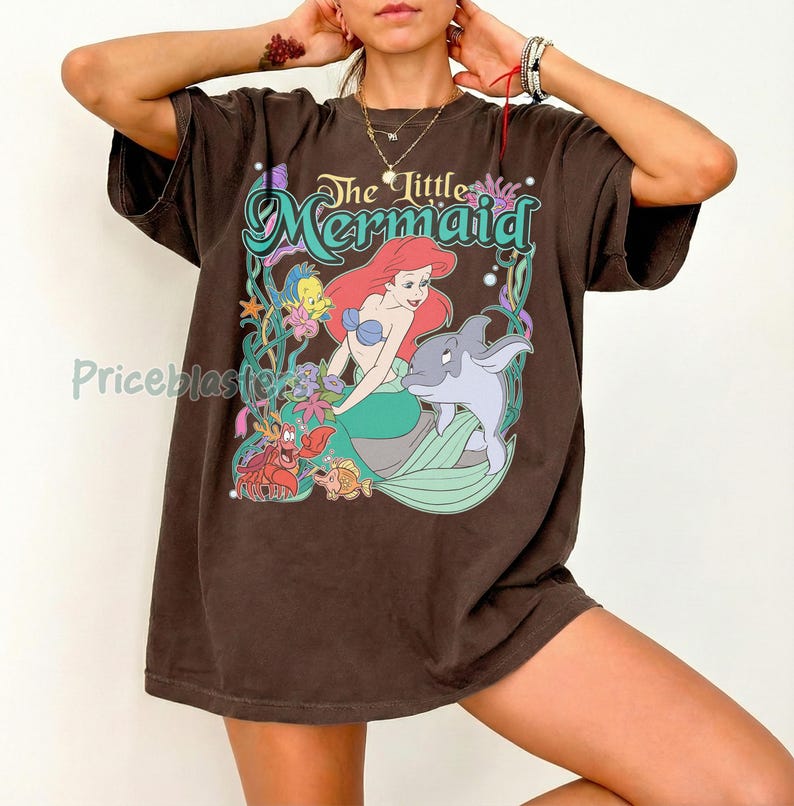 May include: Brown t-shirt featuring a graphic of Ariel from The Little Mermaid. The design includes Ariel with red hair, a blue top, and a green tail, along with a dolphin and other sea creatures. The text "The Little Mermaid" is arched above the image.