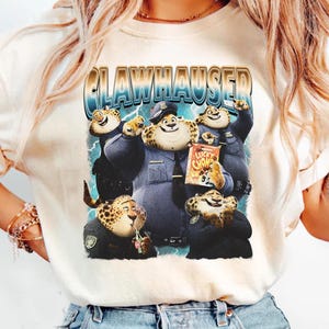 Puede incluir: Camiseta color crema con un gr&aacute;fico de dibujos animados de personajes parecidos a guepardos con uniformes de polic&iacute;a. El gr&aacute;fico incluye el texto "CLAWHAUSER" en fuente azul y dorada, y una caja de cereales "Lucky Chow".