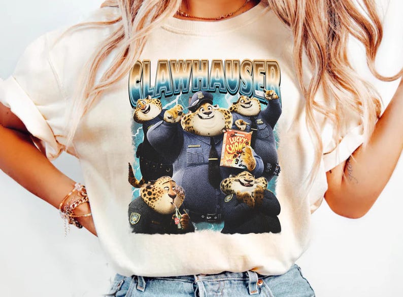 Puede incluir: Camiseta color crema con un gr&aacute;fico de dibujos animados de personajes parecidos a guepardos con uniformes de polic&iacute;a. El gr&aacute;fico incluye el texto "CLAWHAUSER" en fuente azul y dorada, y una caja de cereales "Lucky Chow".