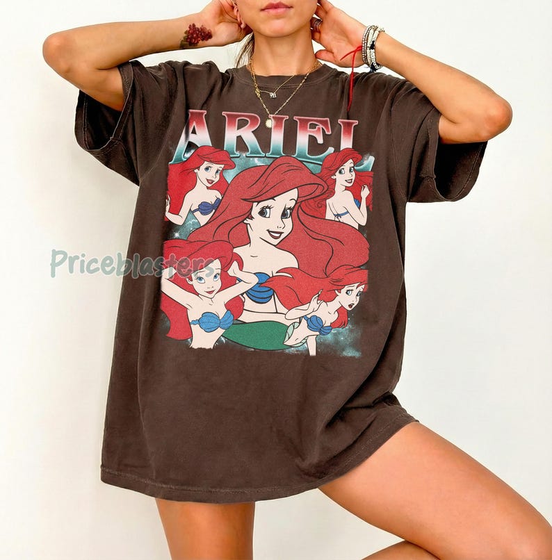 Pu&ograve; includere: Una maglietta marrone con la scritta "ARIEL" in rosso e bianco, e una grafica di Ariel de La Sirenetta. La grafica mostra Ariel con i capelli rossi e un reggiseno a conchiglia blu. La maglietta ha maniche corte e una vestibilit&agrave; ampia.