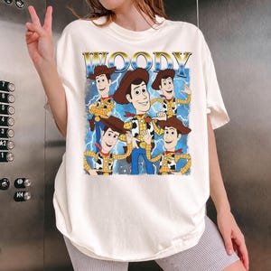 May include: Off-white t-shirt featuring a graphic of Woody from Toy Story. The design includes multiple images of Woody with the word "WOODY" in gold lettering above. The shirt has a relaxed fit.