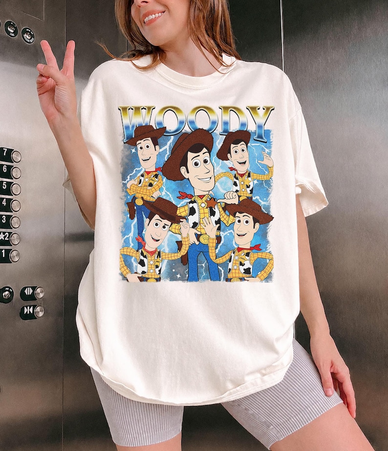 May include: Off-white t-shirt featuring a graphic of Woody from Toy Story. The design includes multiple images of Woody with the word "WOODY" in gold lettering above. The shirt has a relaxed fit.