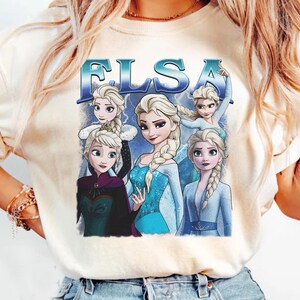 May include: Cream-colored t-shirt featuring a graphic of Elsa from the movie Frozen. The design includes multiple images of Elsa with the word "ELSA" in blue letters. The shirt is a casual wear item.
