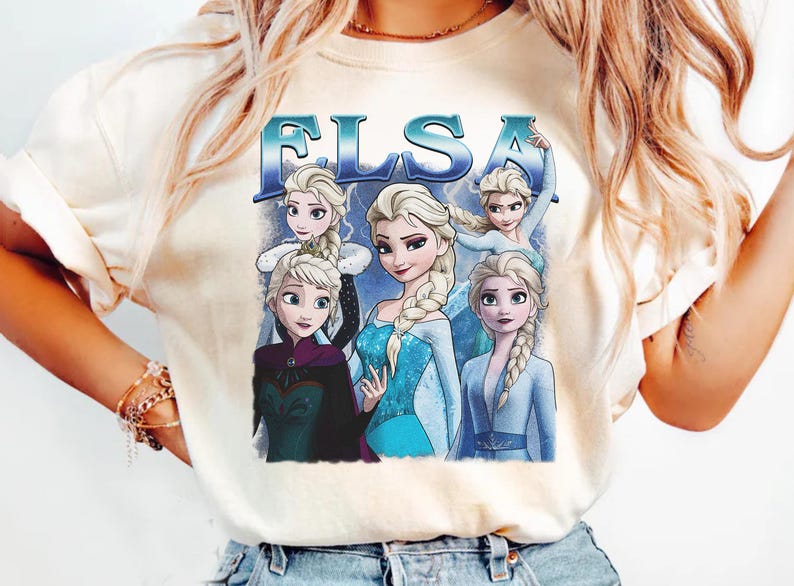 May include: Cream-colored t-shirt featuring a graphic of Elsa from the movie Frozen. The design includes multiple images of Elsa with the word "ELSA" in blue letters. The shirt is a casual wear item.