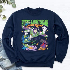May include: Navy blue sweatshirt featuring a graphic of Buzz Lightyear in a dynamic pose, with the text "Buzz Lightyear Laser Blast." The design includes aliens, planets, and space-themed elements, creating a vibrant and playful aesthetic. The sweatshirt is a crew neck style.
