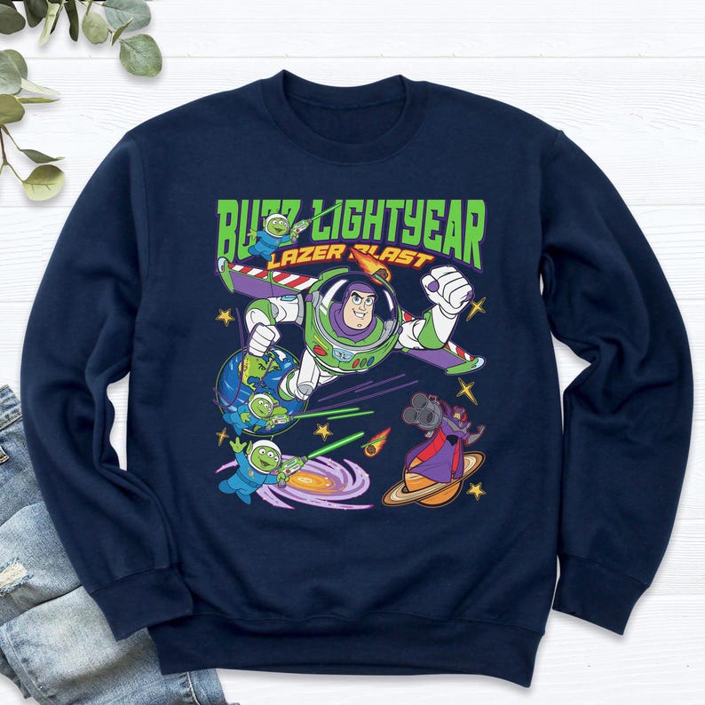 May include: Navy blue sweatshirt featuring a graphic of Buzz Lightyear in a dynamic pose, with the text "Buzz Lightyear Laser Blast." The design includes aliens, planets, and space-themed elements, creating a vibrant and playful aesthetic. The sweatshirt is a crew neck style.