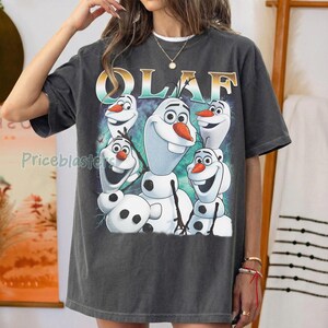 Puede incluir: Camiseta gris oscuro con un gr&aacute;fico de Olaf de Frozen. El dise&ntilde;o incluye m&uacute;ltiples personajes de Olaf con narices de zanahoria y brazos de palo. La palabra "OLAF" est&aacute; impresa en oro y turquesa.