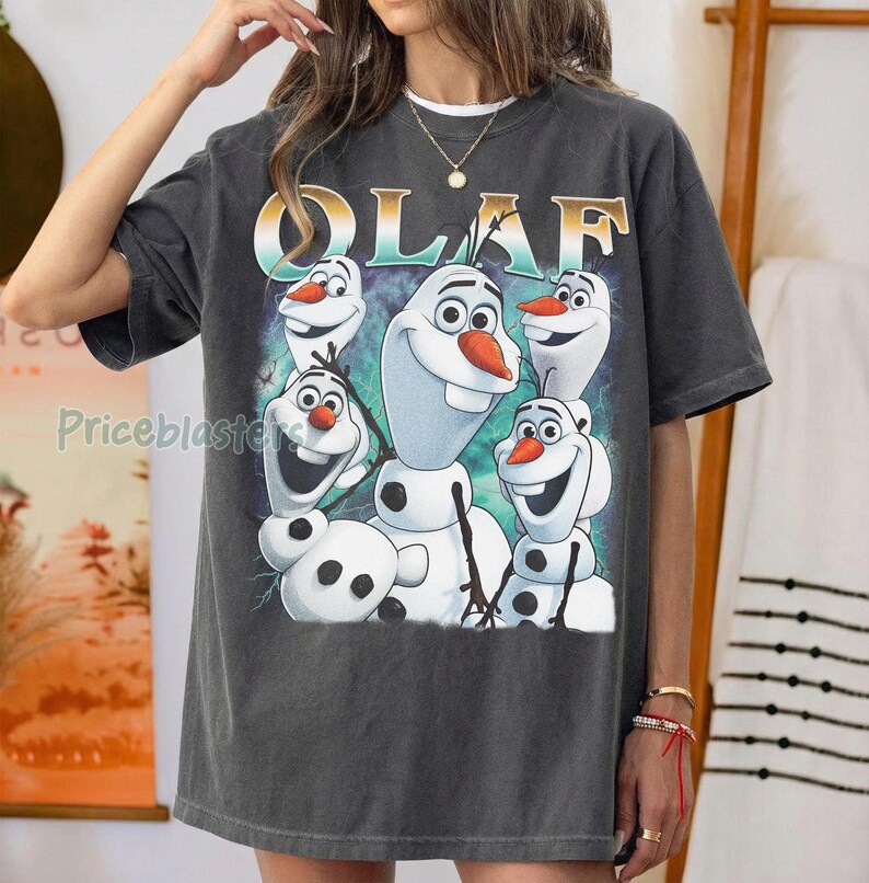 Puede incluir: Camiseta gris oscuro con un gr&aacute;fico de Olaf de Frozen. El dise&ntilde;o incluye m&uacute;ltiples personajes de Olaf con narices de zanahoria y brazos de palo. La palabra "OLAF" est&aacute; impresa en oro y turquesa.