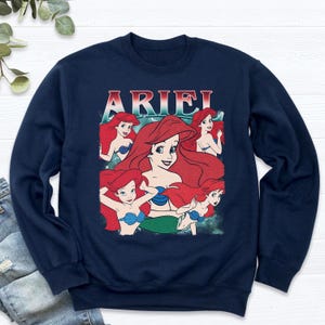 Pu&ograve; includere: Felpa blu navy con una grafica di Ariel de La Sirenetta. Il design include pi&ugrave; immagini di Ariel con capelli rossi e un reggiseno a conchiglia blu. La parola "ARIEL" &egrave; stampata in alto in un font retr&ograve;.
