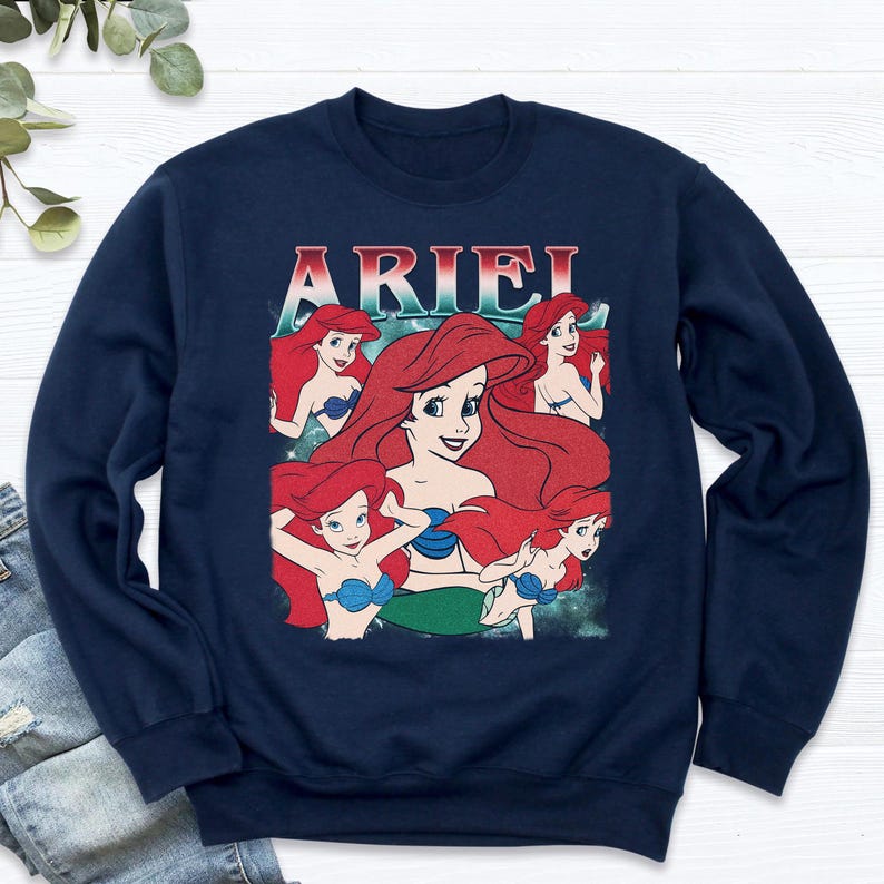 Pu&ograve; includere: Felpa blu navy con una grafica di Ariel de La Sirenetta. Il design include pi&ugrave; immagini di Ariel con capelli rossi e un reggiseno a conchiglia blu. La parola "ARIEL" &egrave; stampata in alto in un font retr&ograve;.