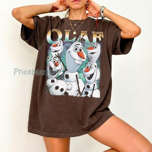 Puede incluir: Camiseta marr&oacute;n con la palabra "OLAF" en letras doradas y un dibujo de Olaf el mu&ntilde;eco de nieve de Frozen. El dibujo muestra a Olaf con una expresi&oacute;n sonriente y una nariz de zanahoria. La camiseta tiene mangas cortas.