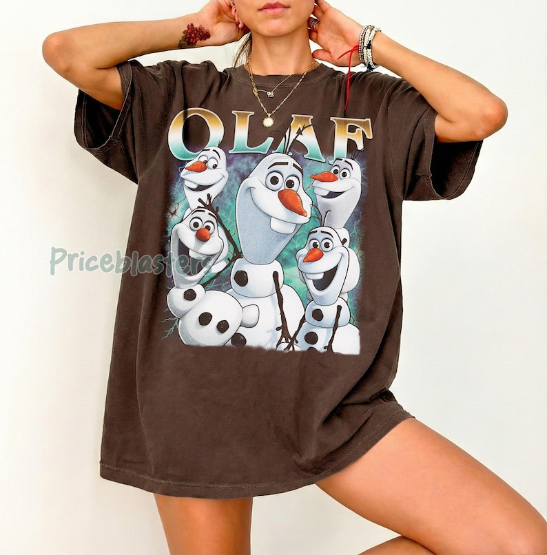 Puede incluir: Camiseta marr&oacute;n con la palabra "OLAF" en letras doradas y un dibujo de Olaf el mu&ntilde;eco de nieve de Frozen. El dibujo muestra a Olaf con una expresi&oacute;n sonriente y una nariz de zanahoria. La camiseta tiene mangas cortas.