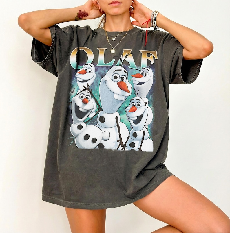 Puede incluir: Camiseta gris oscuro con la palabra "OLAF" en letras doradas sobre un gr&aacute;fico del personaje del mu&ntilde;eco de nieve de la pel&iacute;cula Frozen. El gr&aacute;fico muestra a Olaf en varias poses con un fondo azul y verde.