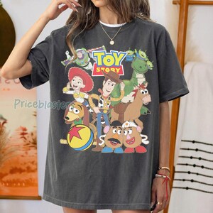 May include: Dark gray t-shirt featuring a colorful graphic of characters from the animated film "Toy Story." The design includes Woody, Buzz Lightyear, Jessie, and other characters, with the "Toy Story" logo at the top. The shirt is a relaxed fit.