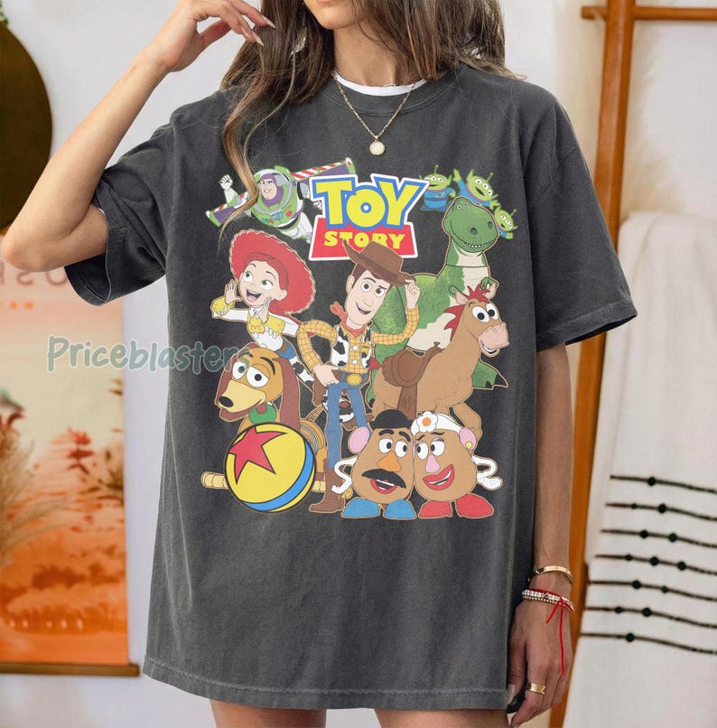 May include: Dark gray t-shirt featuring a colorful graphic of characters from the animated film "Toy Story." The design includes Woody, Buzz Lightyear, Jessie, and other characters, with the "Toy Story" logo at the top. The shirt is a relaxed fit.