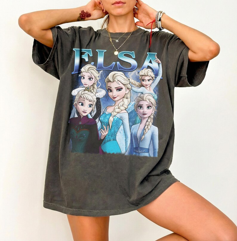 May include: Dark gray t-shirt featuring the name "ELSA" in blue letters above a graphic of the character Elsa from the animated movie Frozen. The graphic shows Elsa in various poses, wearing different outfits.