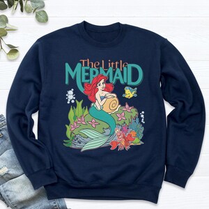May include: Navy blue sweatshirt featuring a colorful graphic of Ariel from The Little Mermaid. The design includes Ariel, a seashell, and other sea creatures. The words "The Little Mermaid" are printed above the image in teal and orange.