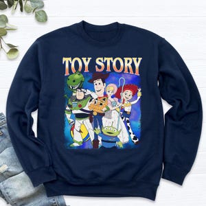 Puede incluir: Sudadera azul marino con un gr&aacute;fico de los personajes de Toy Story, incluyendo a Woody, Buzz Lightyear y Jessie. Las palabras "TOY STORY" est&aacute;n impresas en la parte superior en naranja y blanco. La sudadera es de cuello redondo.