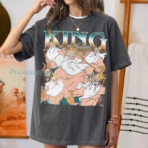 May include: Dark gray t-shirt featuring a graphic of King Triton with a trident. The text "KING Triton" is displayed above the illustration in a teal and gold font. The shirt has a vintage, worn look.