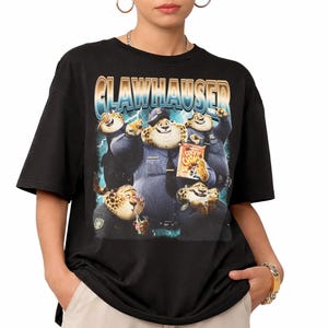 Puede incluir: Camiseta negra con un gr&aacute;fico de Clawhauser de Zootopia. El dise&ntilde;o incluye el texto "CLAWHAUSER" en una fuente retro e im&aacute;genes de Clawhauser con uniforme de polic&iacute;a, sosteniendo bocadillos. La camiseta es de cuello redondo y manga corta.