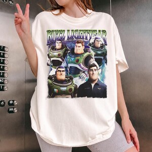 May include: Cream-colored t-shirt featuring a graphic of Buzz Lightyear from the movie. The design includes multiple images of the character and the text "BUZZ LIGHTYEAR" in a purple and green font. The shirt has short sleeves and a relaxed fit.