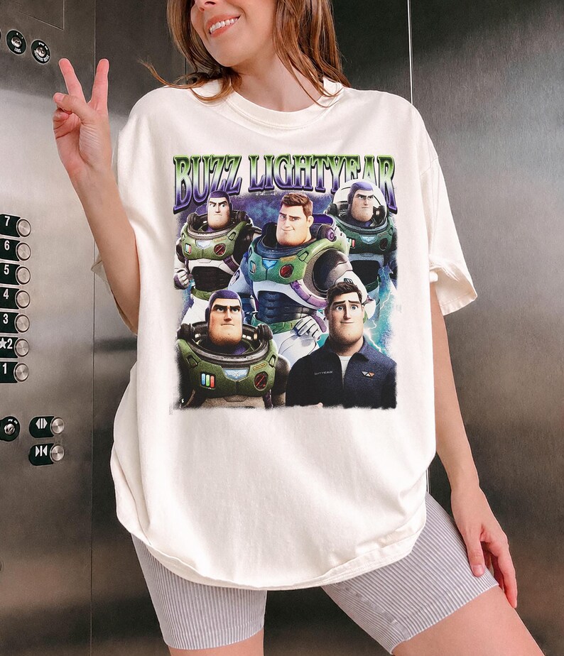 May include: Cream-colored t-shirt featuring a graphic of Buzz Lightyear from the movie. The design includes multiple images of the character and the text "BUZZ LIGHTYEAR" in a purple and green font. The shirt has short sleeves and a relaxed fit.