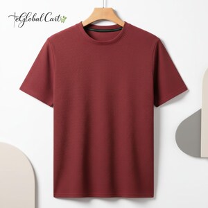 May include: A short-sleeved, burgundy t-shirt with a ribbed texture, hanging on a wooden hanger. The shirt has a crew neck with a green trim. The Global Cart logo is visible in the top left corner.