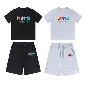 Puede incluir: Un conjunto de camisetas y pantalones cortos negros y grises. Las camisetas y los pantalones cortos tienen la palabra "TRAPSTAR" en colores arcoíris. El texto "IT'S A SECRET" está debajo de la palabra "TRAPSTAR".