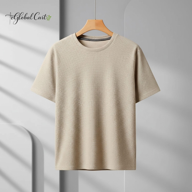 May include: A short-sleeved, beige t-shirt with a textured, ribbed design. The shirt has a crew neck with a dark trim and is hanging on a wooden hanger. The Global Cart logo is visible in the upper left corner.