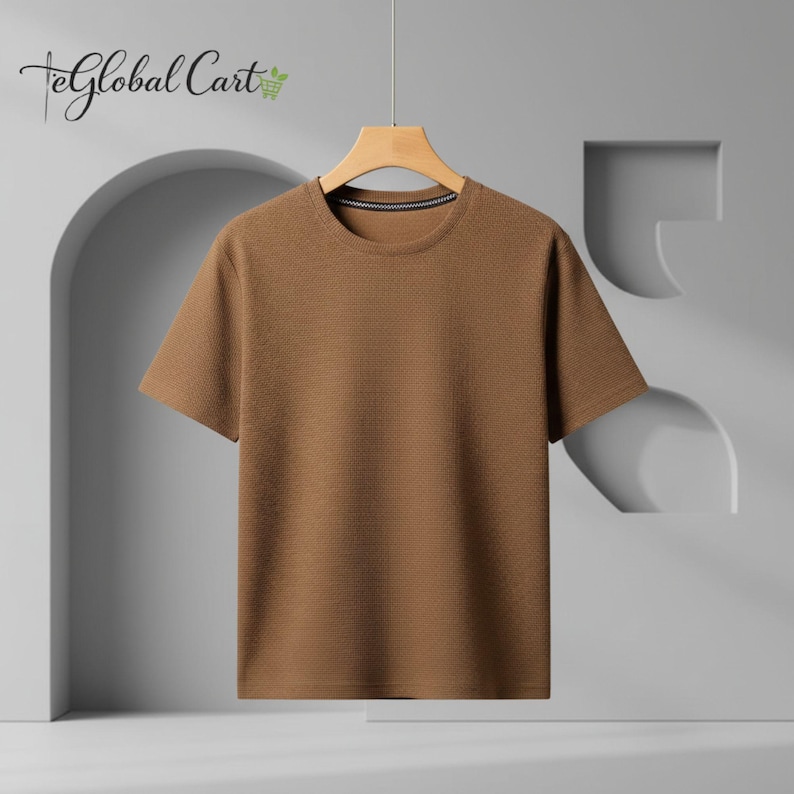 May include: A short-sleeved, brown t-shirt hangs from a wooden hanger. The shirt has a textured pattern and a crew neck with a thin, dark stripe. The background is a light gray with architectural design elements.