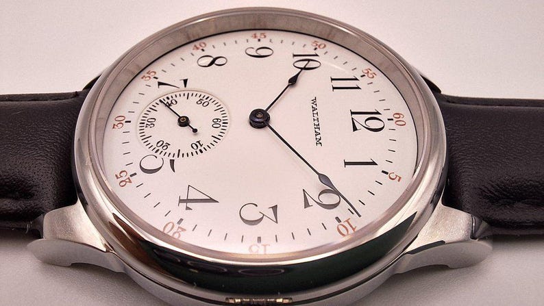 May include: A close-up of a vintage Waltham watch with a white face, black hands, and a small subdial. The watch has a silver-colored case and a black leather band. The watch face features black Arabic numerals and the word "WALTHAM".