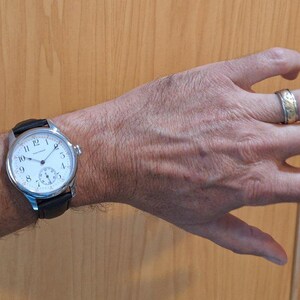 May include: A silver wristwatch with a white face and black leather band is displayed on a person's wrist. The watch has black numerals and a small seconds dial. A gold ring with a decorative pattern is on the person's finger.
