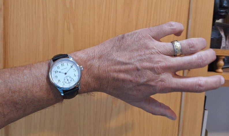 May include: A silver wristwatch with a white face and black leather band is displayed on a person's wrist. The watch has black numerals and a small seconds dial. A gold ring with a decorative pattern is on the person's finger.