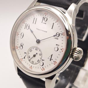 May include: A silver-toned Waltham wristwatch with a white face and black leather band. The watch features black numerals, a small seconds dial, and a textured crown. The watch face displays the brand name.
