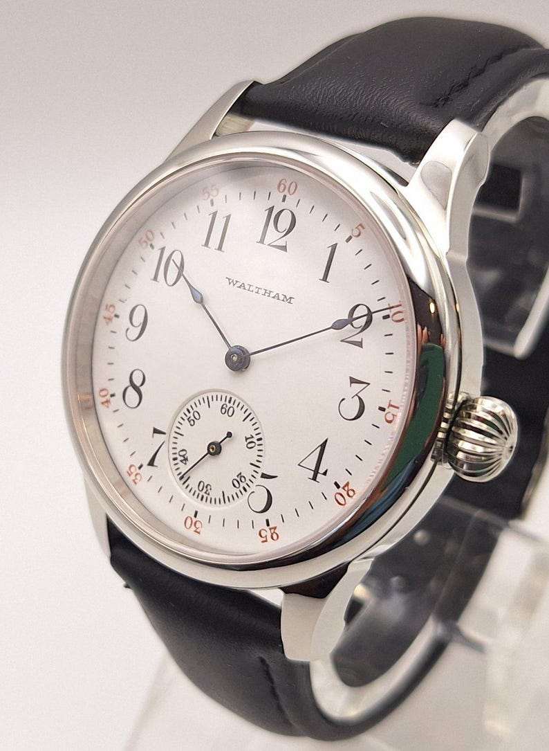 May include: A silver-toned Waltham wristwatch with a white face and black leather band. The watch features black numerals, a small seconds dial, and a textured crown. The watch face displays the brand name.