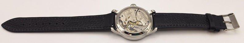 May include: A silver-toned wristwatch with a black leather band. The watch face reveals the intricate internal mechanisms. The band is secured with a silver buckle. The watch is a classic accessory.